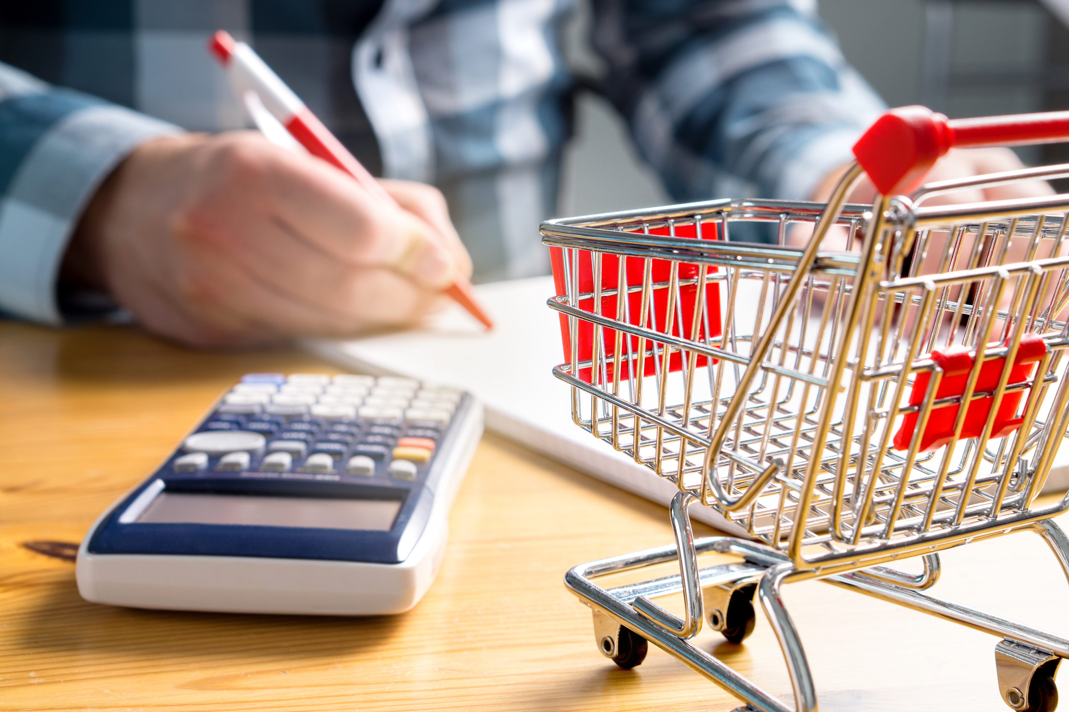 Hand holding a pen on a notepad, with a calculator and a mini shopping cart on a table.