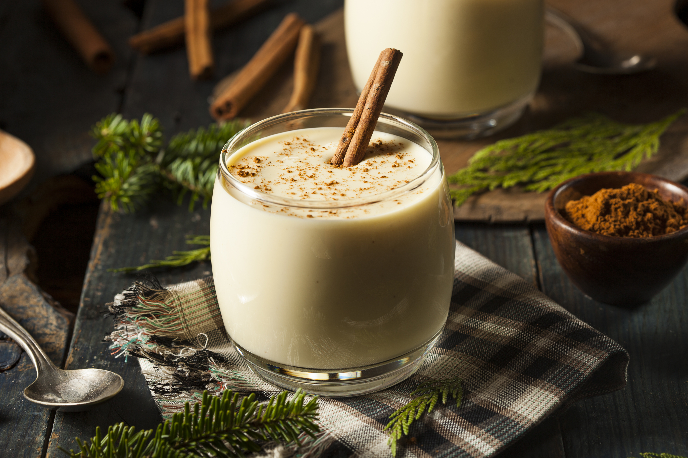 Glass of eggnog next to a bowl of ground cinnamon on a table decorated with pine tree branches and plaid napkins.