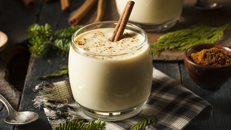 Un verre de lait de poule et un bol de cannelle moulue sur une table décorée de branches de pin et de serviettes à carreaux.