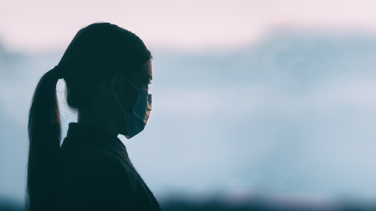 Une jeune femme portant un masque médical regarde au loin, arrière-plan flou.