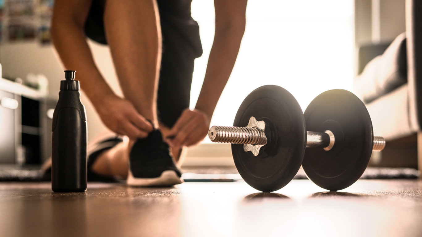 Person kneeling down and tying their shoes in their home gym, with weights and a water bottle nearby. 