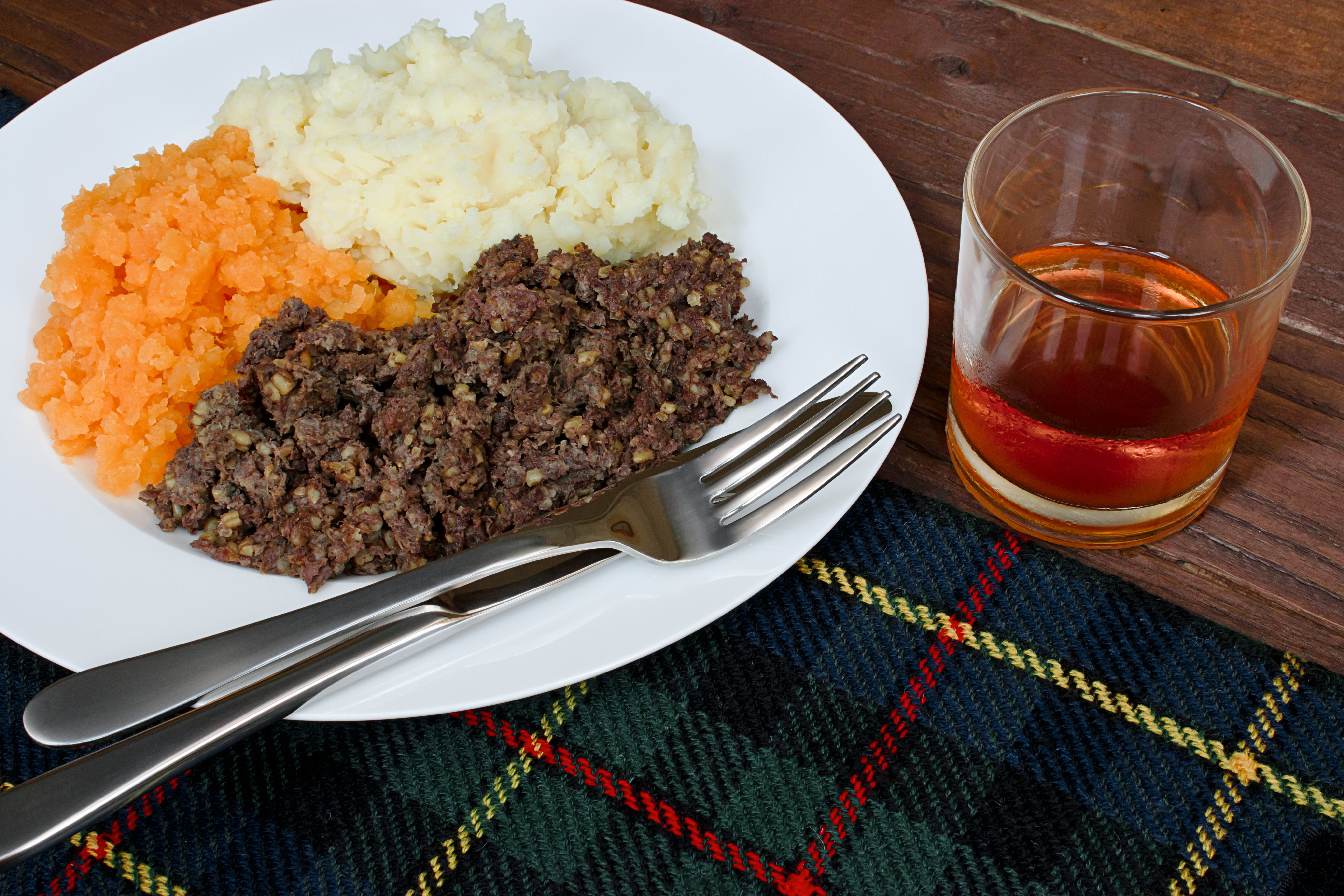 A plate of haggis, mashed potatoes and turnips with a glass of Scotch whisky placed on a Scottish tartan tablecloth.