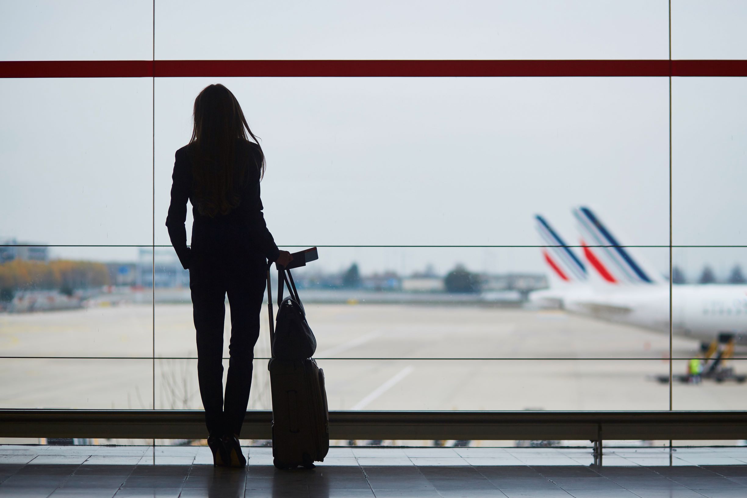 Woman holding suitcase in an airport looking out the window at airplanes on tarmac.