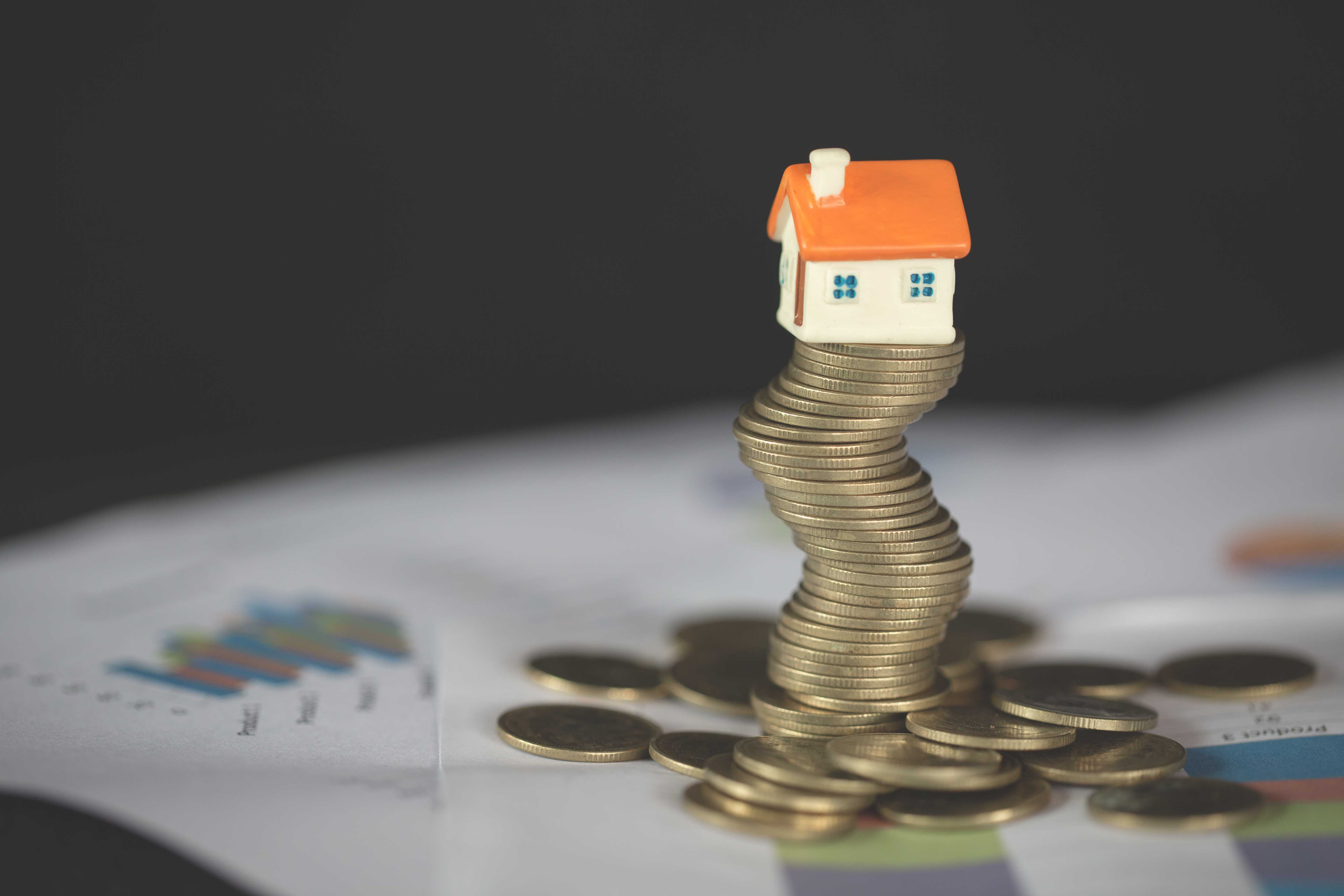 A miniature house made of painted clay, perched on a pile of coins, placed on financial documents. 