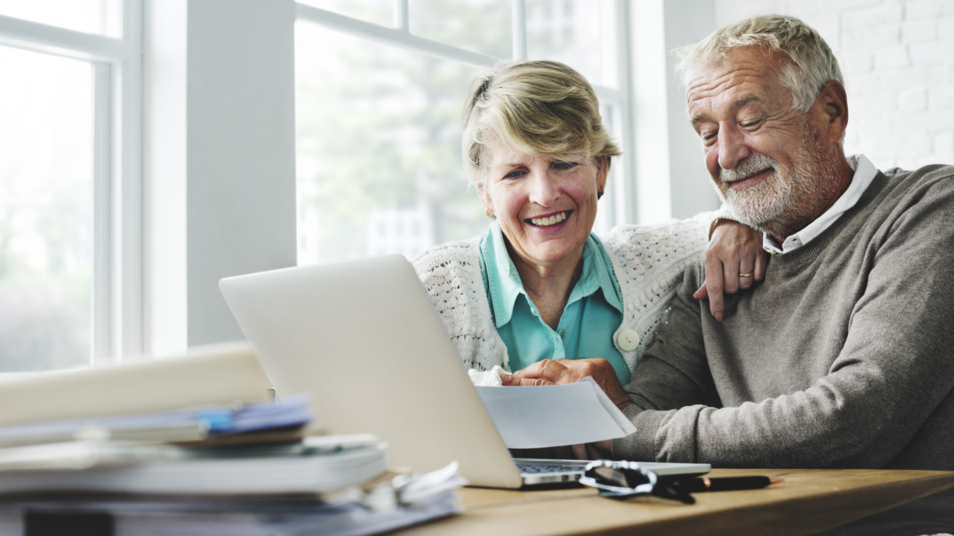 An older couple looking at a laptop and a piece of paper while sitting at a table.
