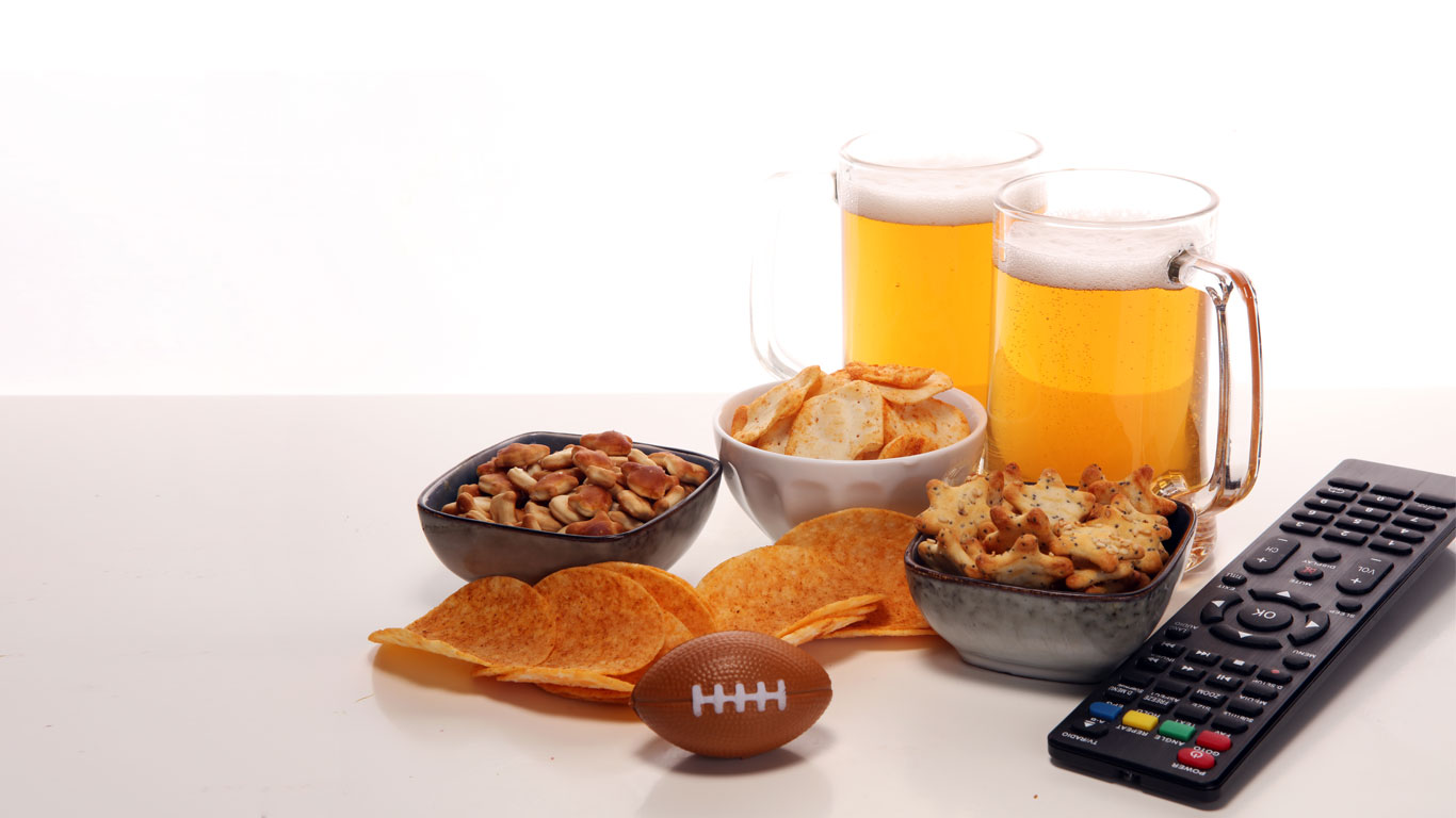 A miniature football next to a TV remote, snacks and two frothy ice-cold mugs of beer