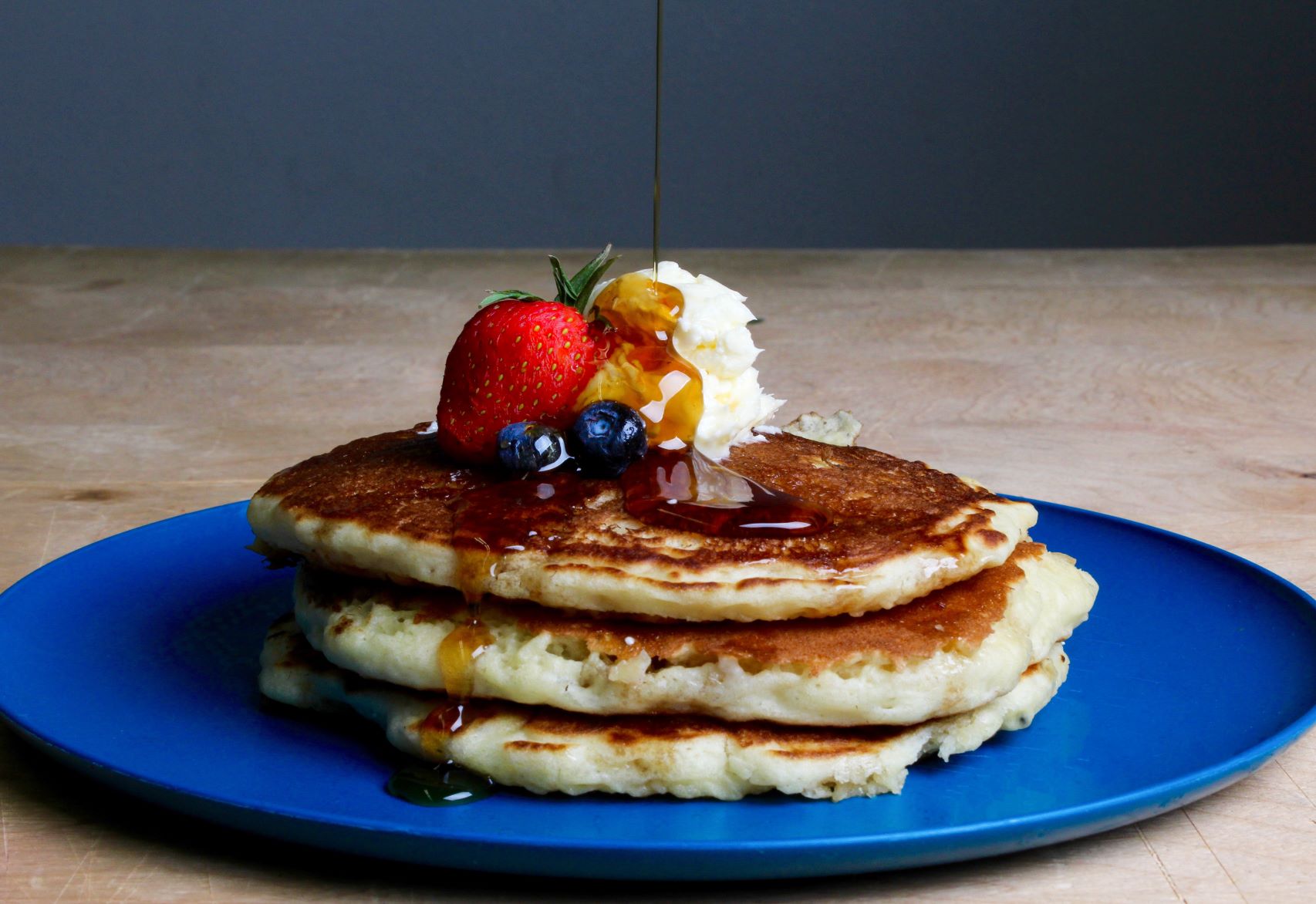A pile of three pancakes on a blue plate with maple syrup, berries and butter.