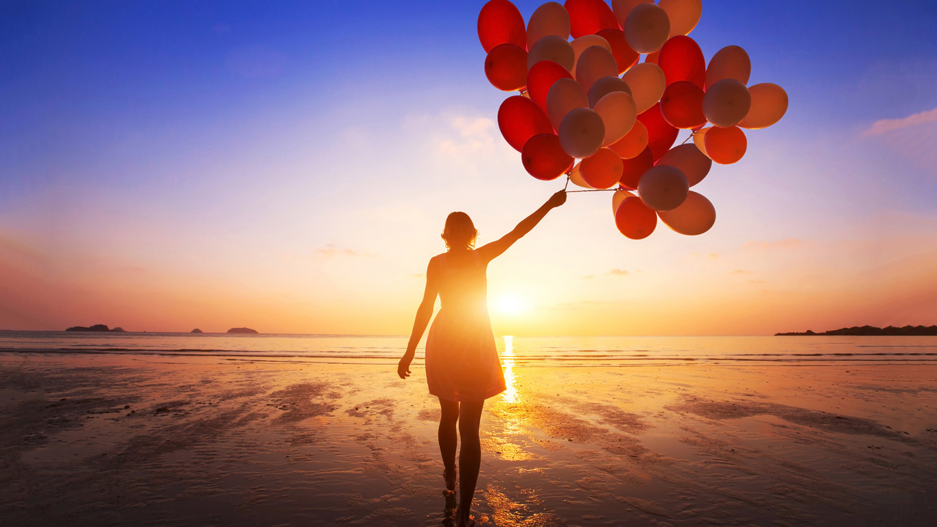A young woman holding a bunch of red balloons on a beach at sunrise.