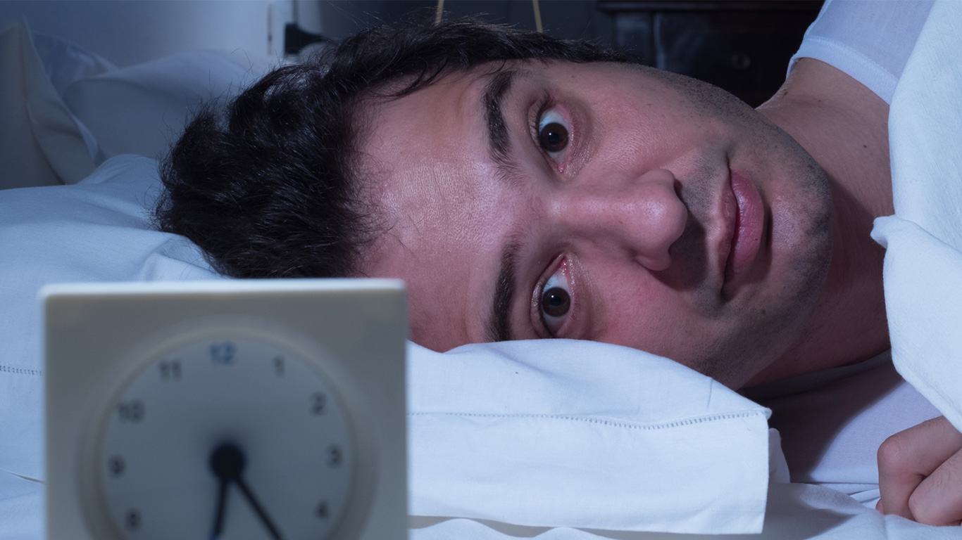 A man lying in bed with bleary eyes staring at an alarm clock.