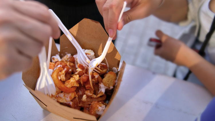 Des mains qui piquent des fourchettes dans un plat de poutine pour emporter posé sur une table 